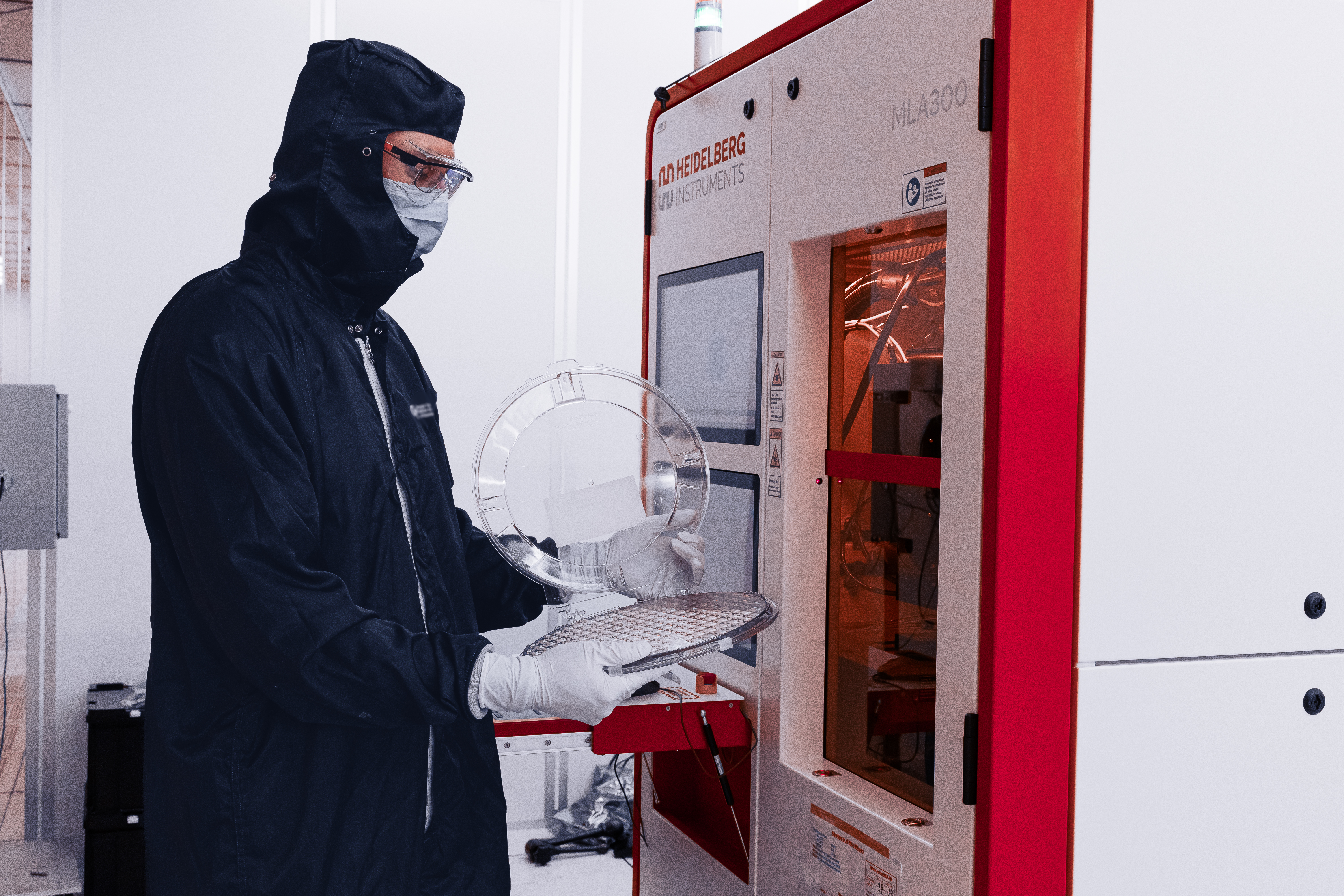 A researcher wearing cleanroom protective gear loads or unloads a semiconductor wafer carrier at a Heidelberg Instruments lithography system in a laboratory cleanroom.
