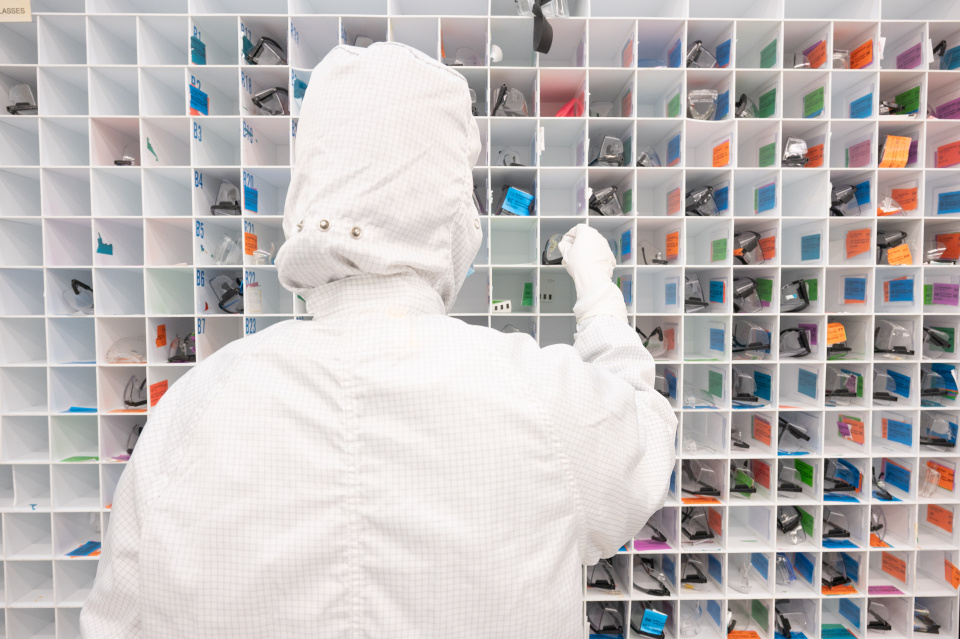 Close-up of hands assembling and testing a microelectronics circuit on breadboards with wires, probes, and a small semiconductor device in a laboratory setting.  A person wearing a cleanroom suit selects labeled components from a wall of organized storage compartments in a microelectronics fabrication facility.