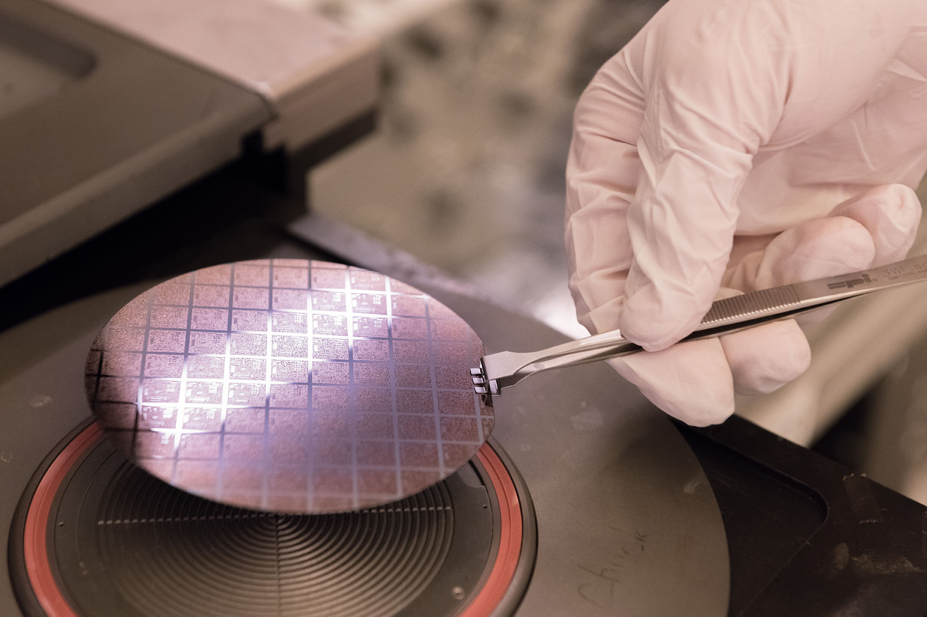 Gloved hands use precision tweezers to handle a semiconductor wafer with patterned microchips during fabrication or inspection in a cleanroom environment.