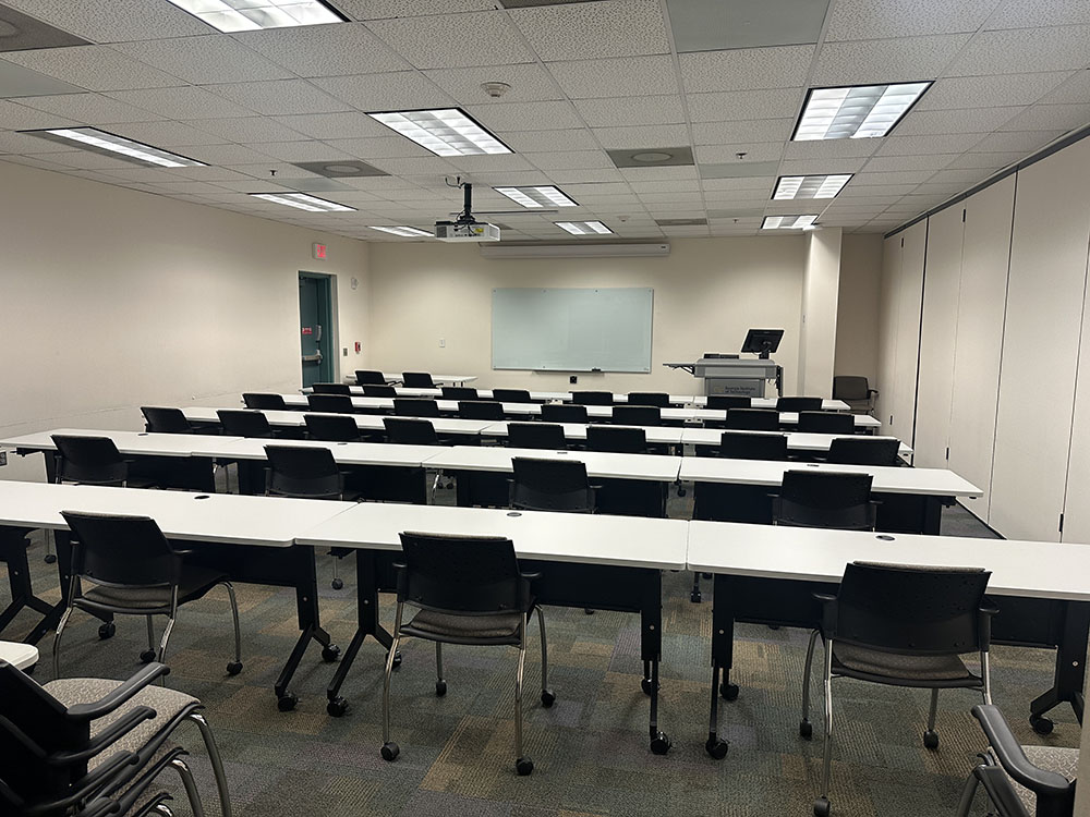 Classroom with rows of white tables and black chairs facing a whiteboard and lectern with a computer and ceiling-mounted projector.