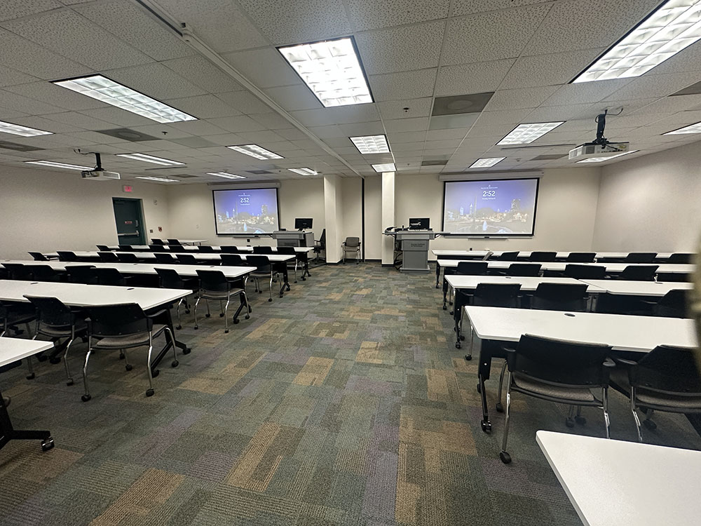 Empty classroom with rows of white tables and black chairs facing two projection screens and instructor podiums at the front, under fluorescent ceiling lights.