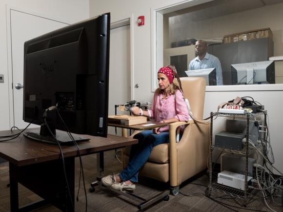 A person seated in a beige chair using a computer setup with multiple cables and devices, facing a large monitor in a testing or research room, with another individual visible through a window in an adjacent control room.