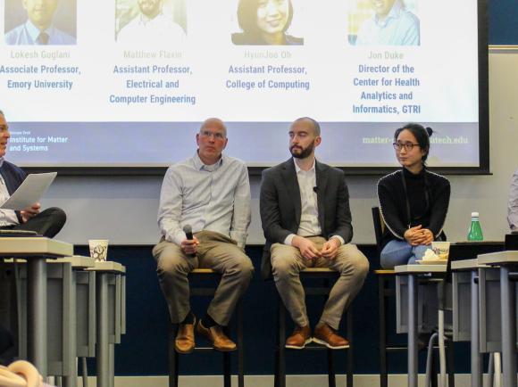 A panel of five speakers sits on tall stools at the front of a classroom, participating in a moderated discussion. The moderator on the left holds papers while addressing the group. A large presentation slide behind the panel displays names and academic titles. Audience members are partially visible in the foreground, and tables, chairs, and a water bottle are arranged throughout the room.