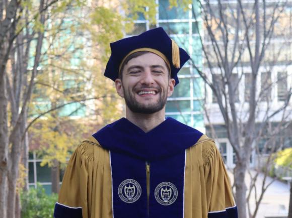 A smiling graduate stands on a tree-lined campus walkway covered with fallen leaves. He is wearing a gold doctoral gown with blue velvet panels and sleeve bars, along with a matching blue tam and tassel. Campus buildings and autumn trees are visible in the background.