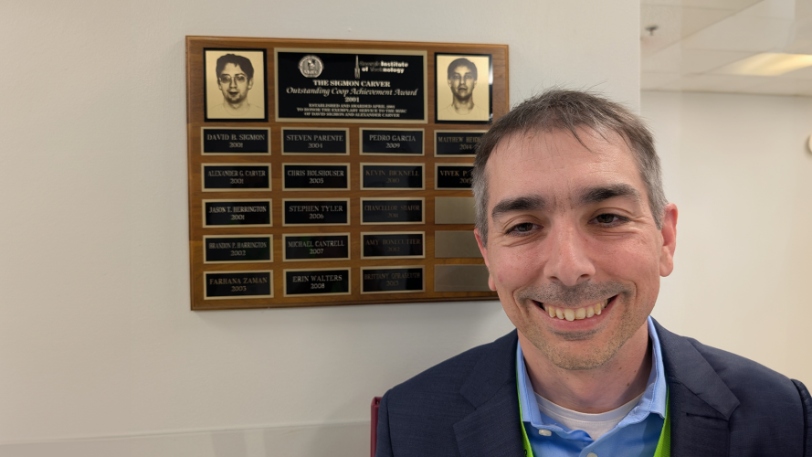 A smiling man in a blazer stands indoors next to a wall-mounted plaque displaying “The Sigmon Carver Outstanding Co-Op Achievement Award” and a list of recipients.