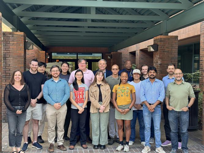 A group of students, researchers, and staff stand together for a group photo outside a brick building beneath a covered walkway.