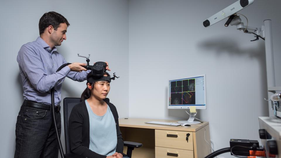 Researcher adjusting a device on another person’s head in a lab, with a computer displaying brain imaging data and a mounted camera in the background.