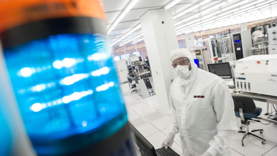 A cleanroom technician in protective gear works at a computer workstation in a semiconductor lab, with a blue signal light in the foreground and lab equipment behind them.