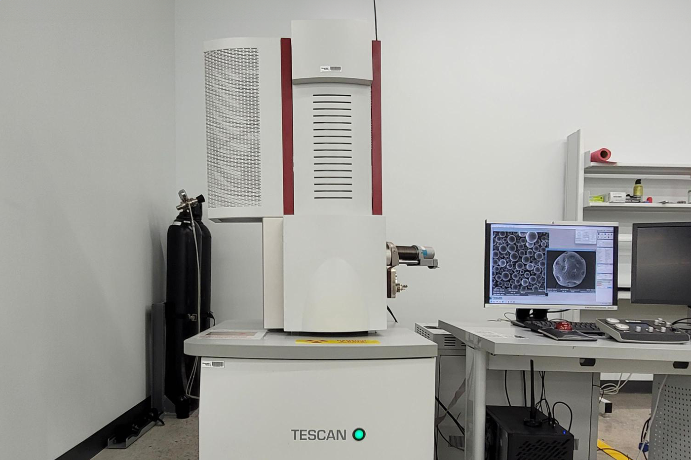 A white TESCAN scanning electron microscope (SEM) stands in a clean laboratory next to gas cylinders and a computer workstation. The monitor displays magnified circular microstructures captured by the microscope.